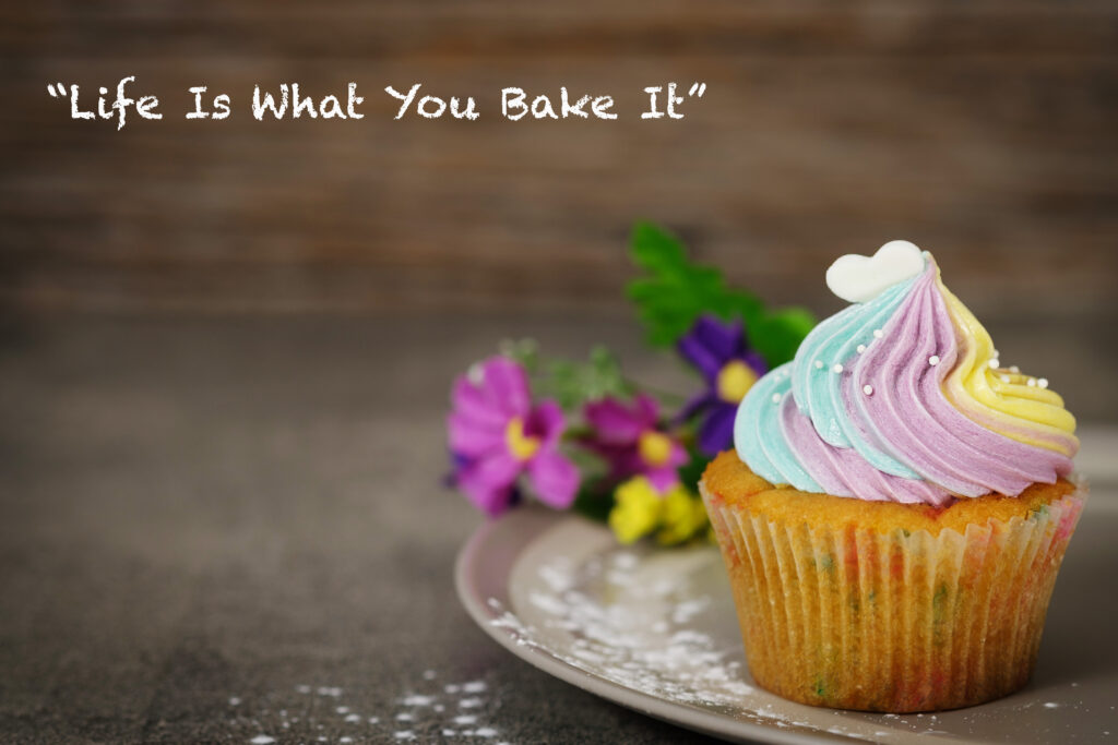 "Life Is What You Bake It" with flowers in the background and cupcake with purple, blue, and yellow frosting.